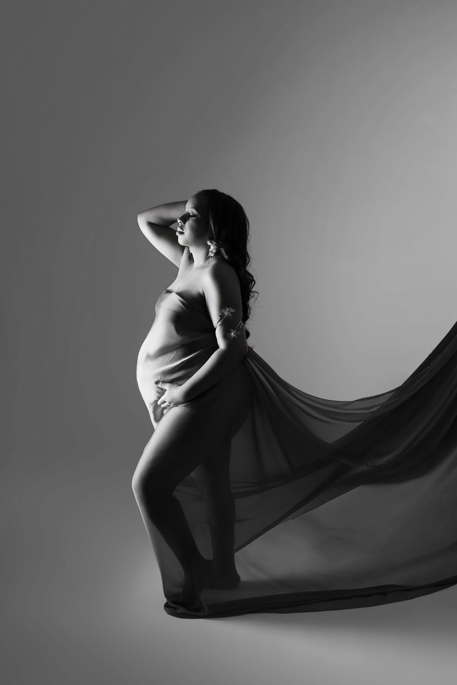 Black and white maternity photo of pregnant woman draped in flowing fabric, posing gracefully in studio.