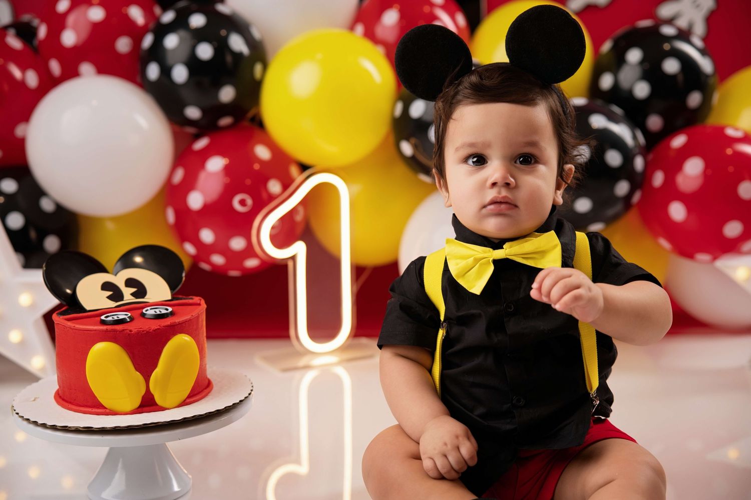 Baby boy in Mickey Mouse outfit celebrates 1st birthday with themed cake and colorful balloons backdrop.