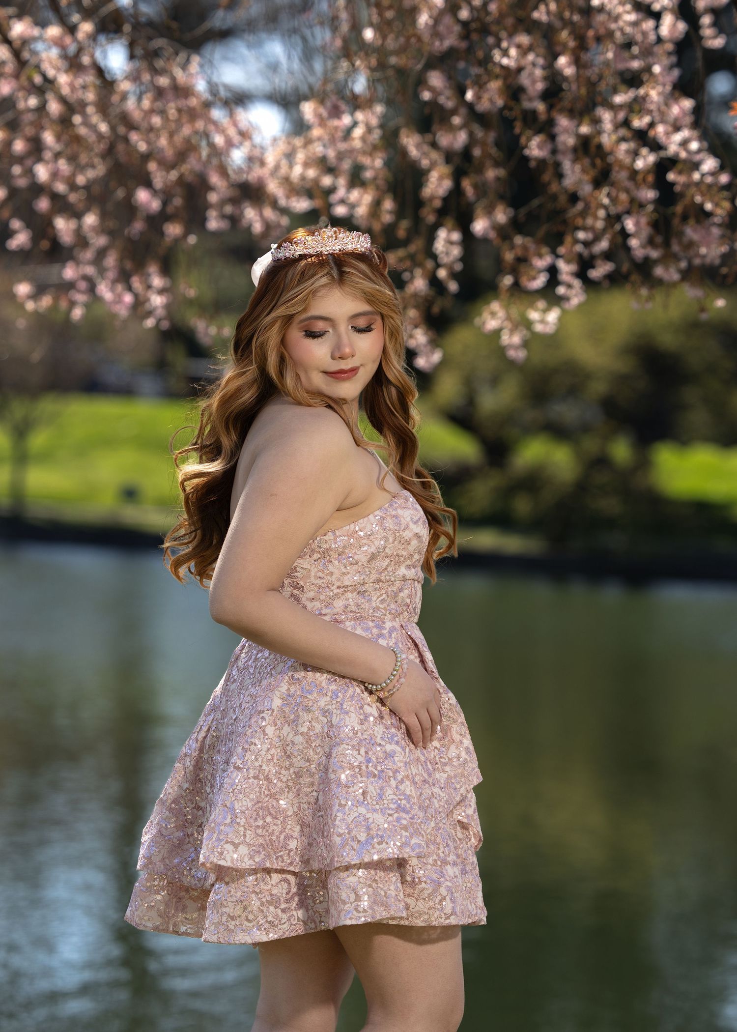 Young woman in pink sequin dress and tiara poses by a pond under blooming cherry blossom trees.
