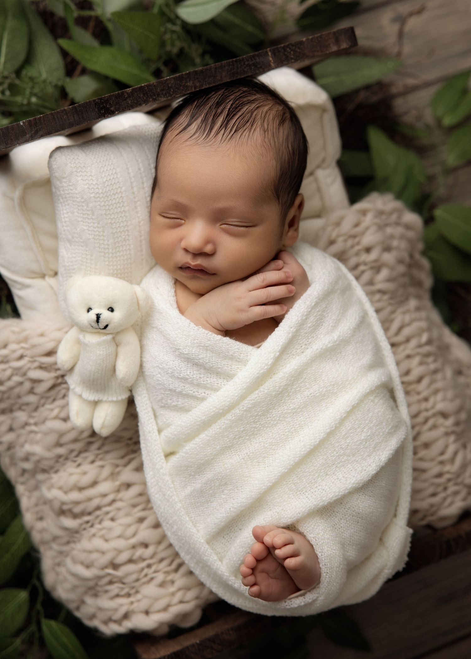 A sleeping infant wrapped in a white blanket lies beside a plush teddy bear on a textured grey surface.