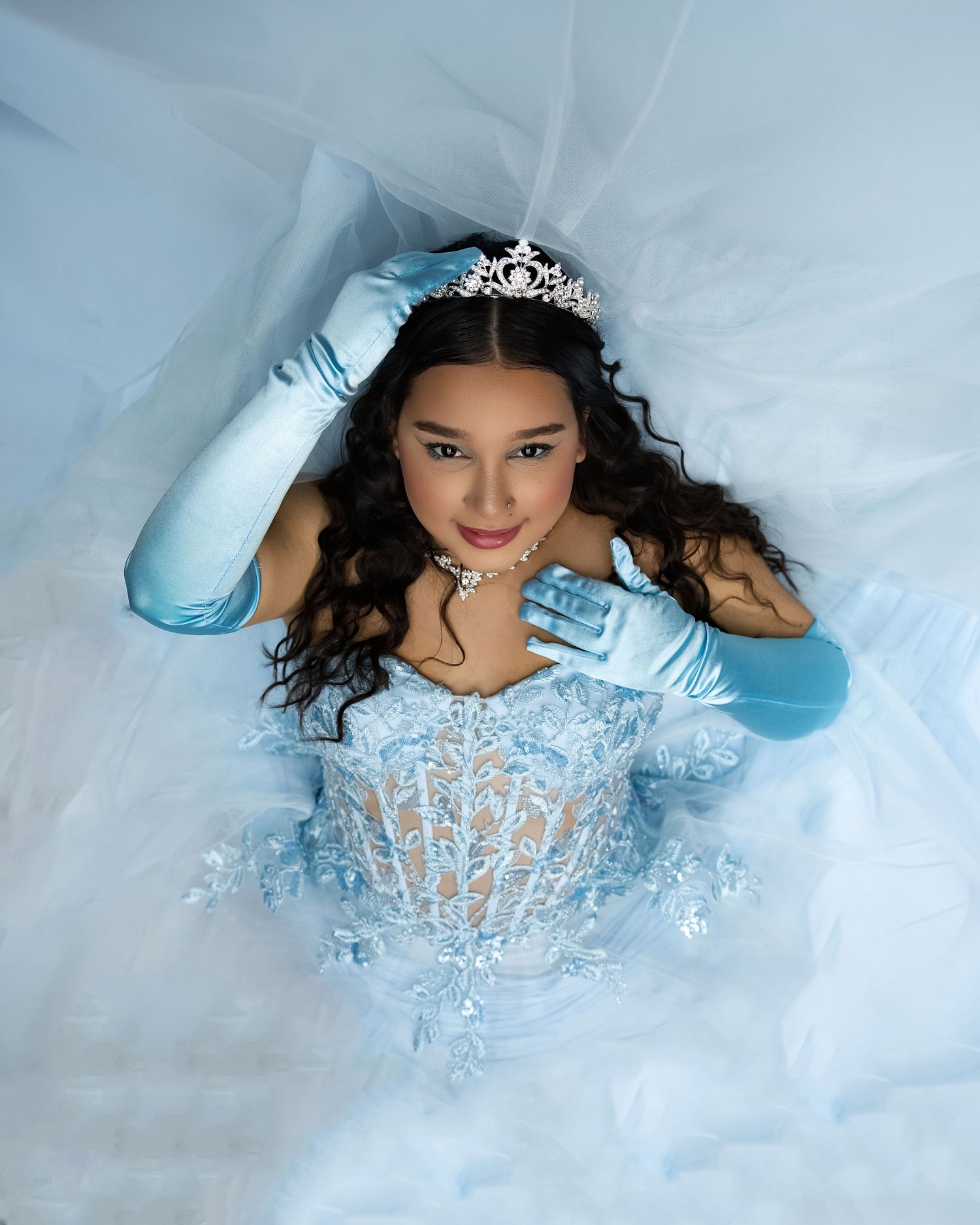 Young woman in blue quinceañera gown with tiara, gloves, and veil poses elegantly for formal portrait.