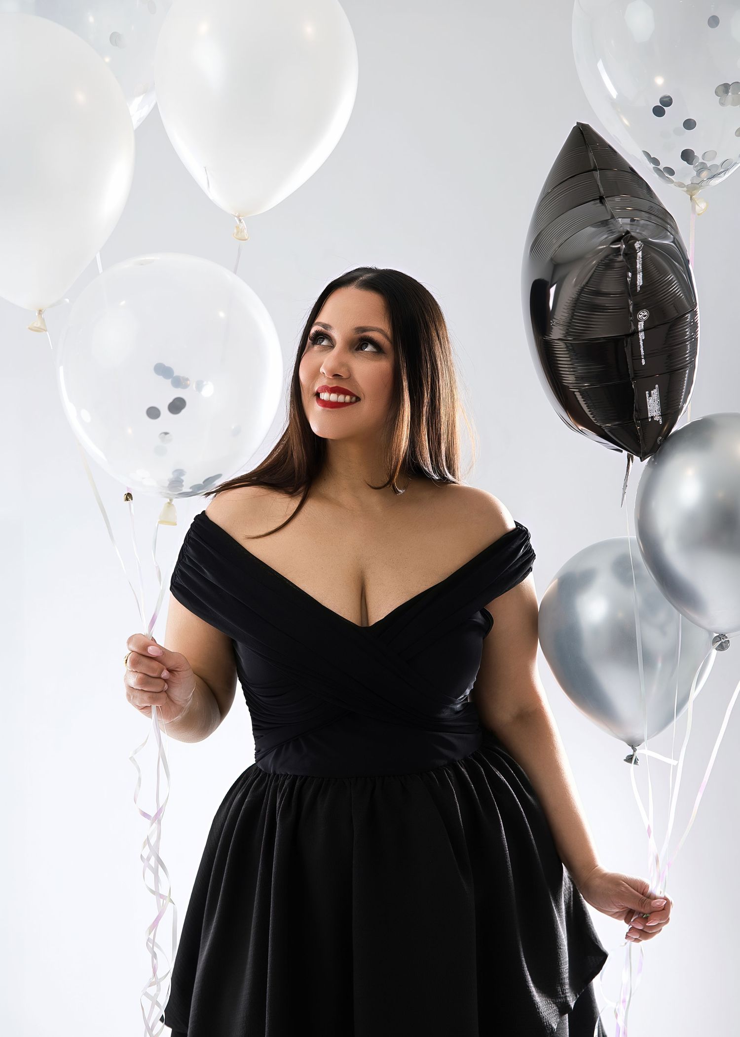 Woman in black off-shoulder dress holding white, silver, and black balloons, smiling against white background.