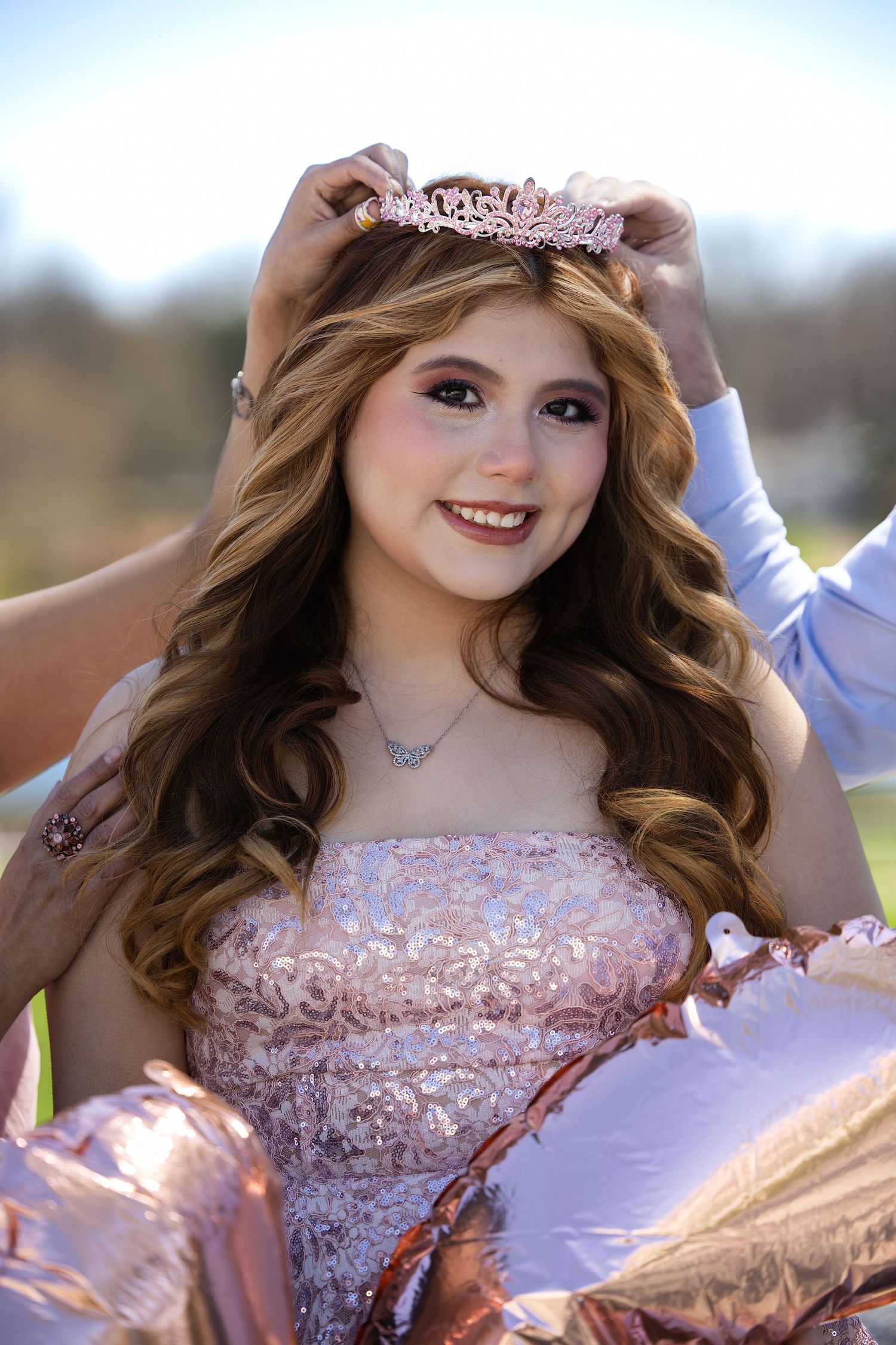 Young girl smiling as a rose gold tiara is placed on her head, wearing a pink gown, quinceañera celebration outdoors.