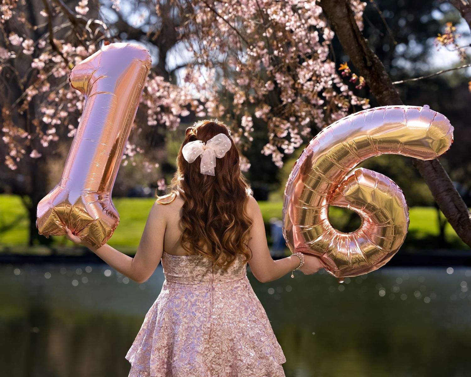 Girl in sparkly dress holding rose gold number 16 balloons outdoors near cherry blossom trees for sweet sixteen.