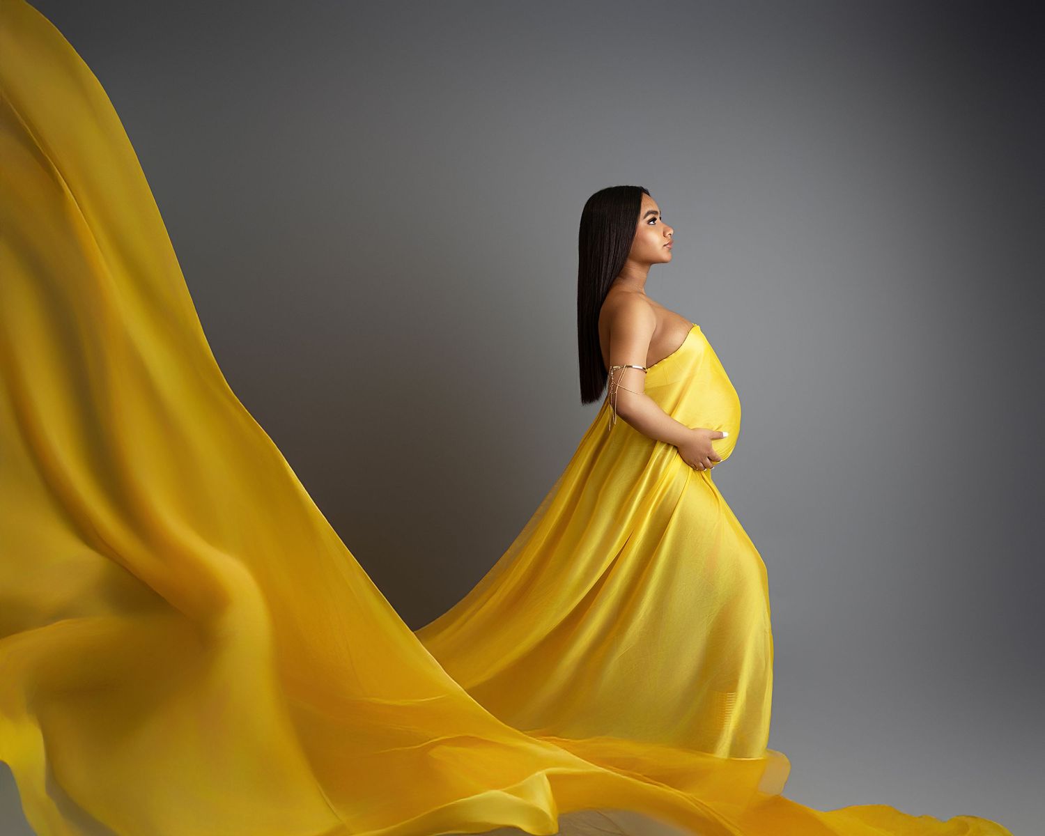 Figure in flowing bright yellow maternity gown poses against grey background as fabric dramatically billows behind.