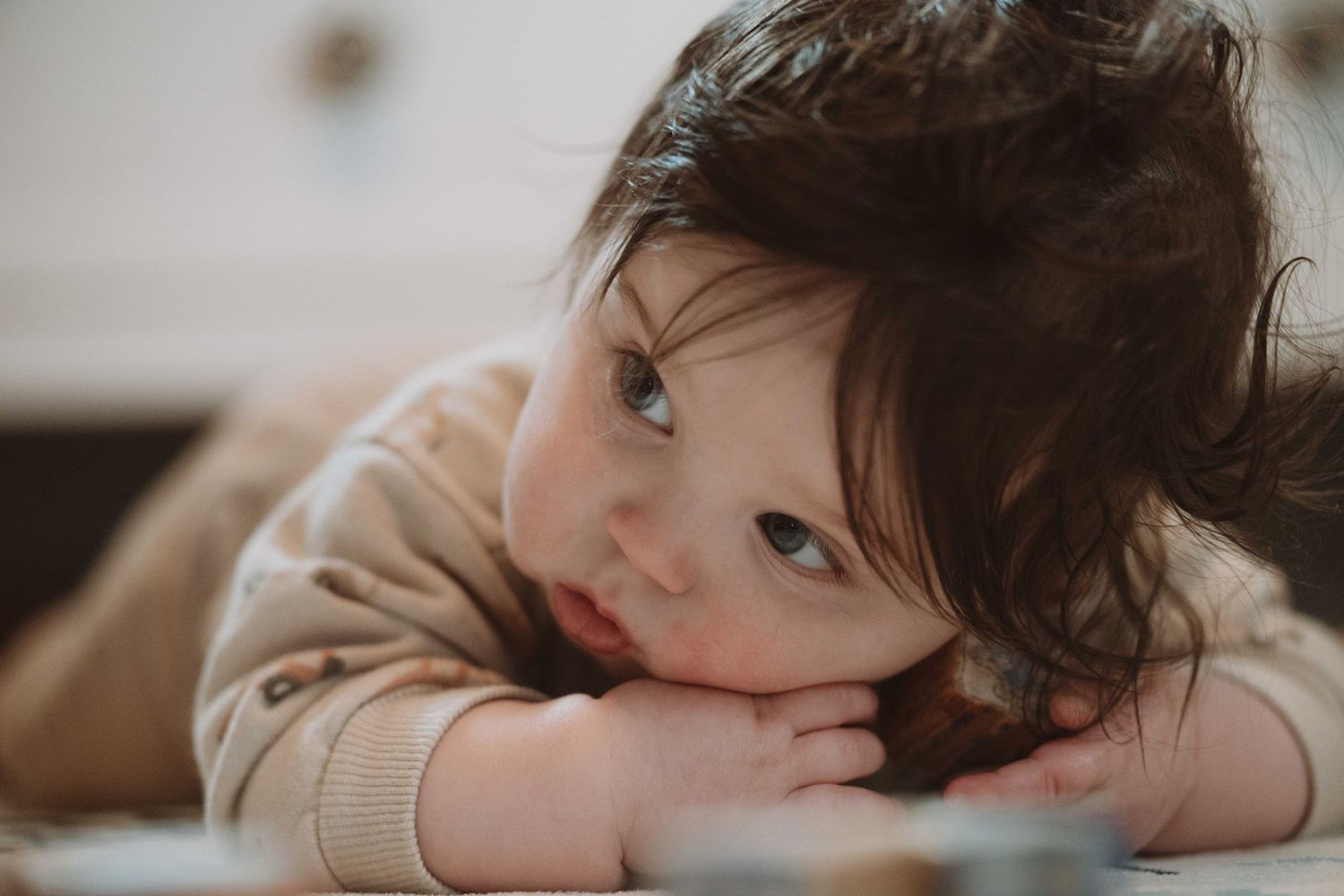 Adorable baby with dark hair resting chin on hands, gazing thoughtfully in a soft, warm-toned portrait photo.