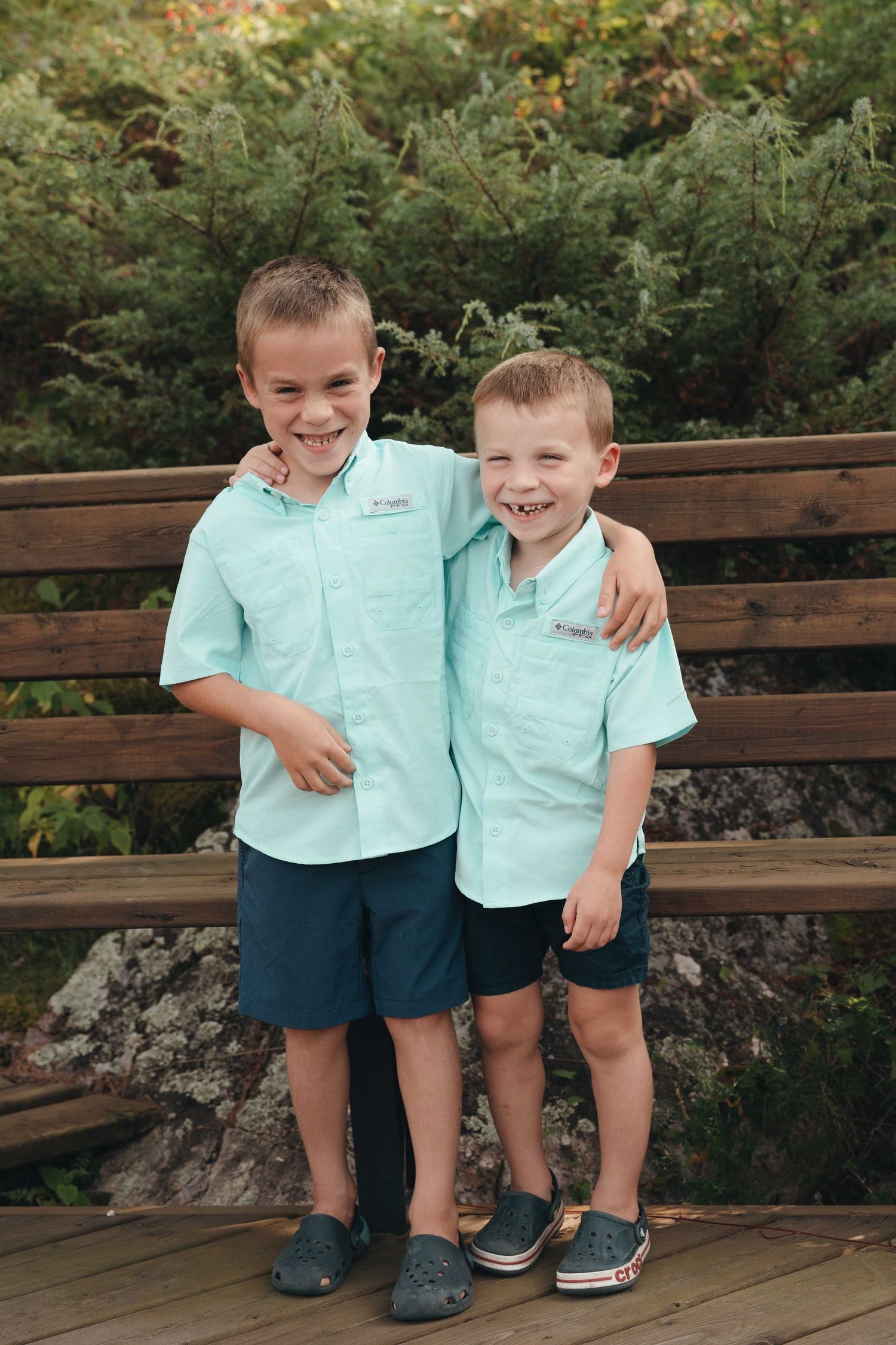 Two young boys in matching mint Columbia shirts and navy shorts smiling together on a wooden deck outdoors.