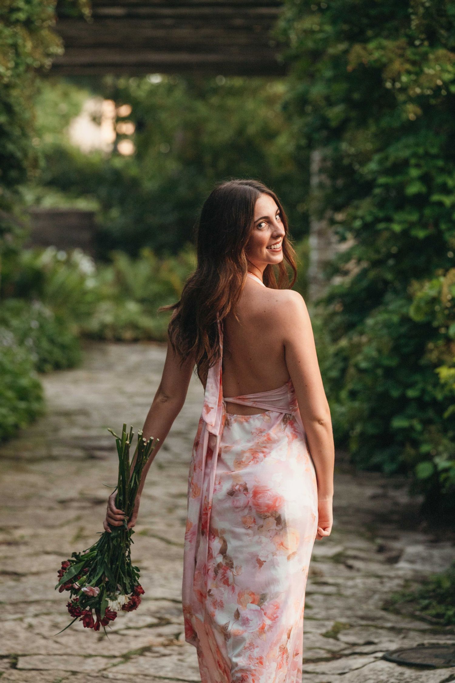 Woman in floral backless dress holding red flowers smiles in lush garden pathway.