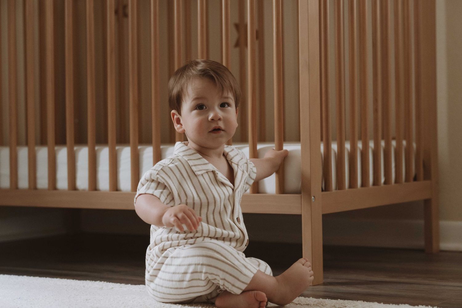 A baby in a striped romper sits on a carpet in front of a wooden crib in a nursery.