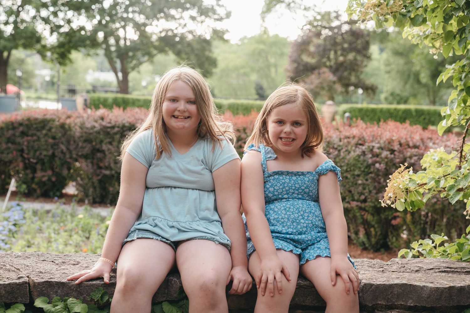 Two smiling blonde girls sitting together on a wooden bench in a lush garden setting.