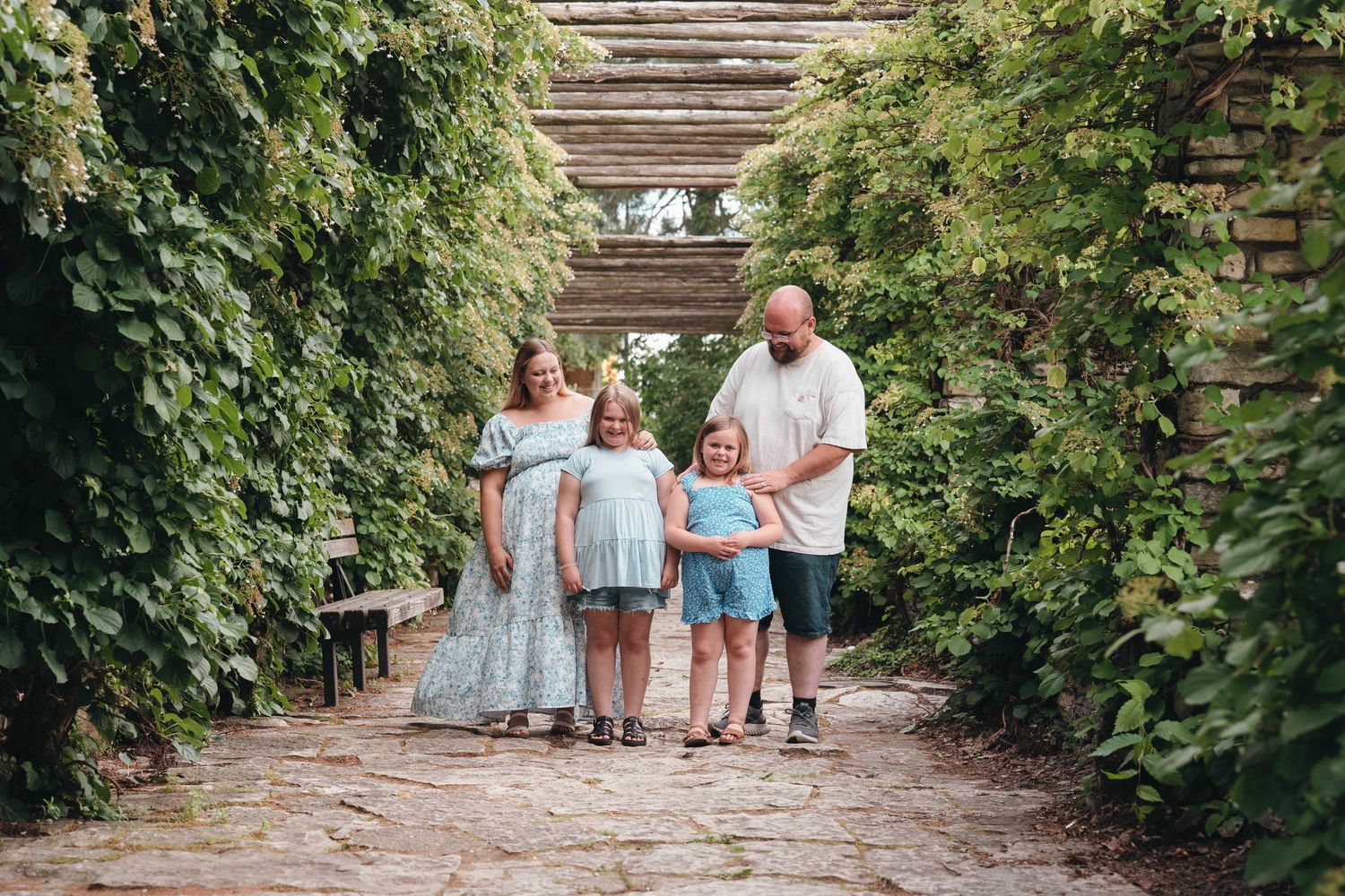 Family of four poses on a stone path beneath a green vine-covered pergola in a lush garden setting.