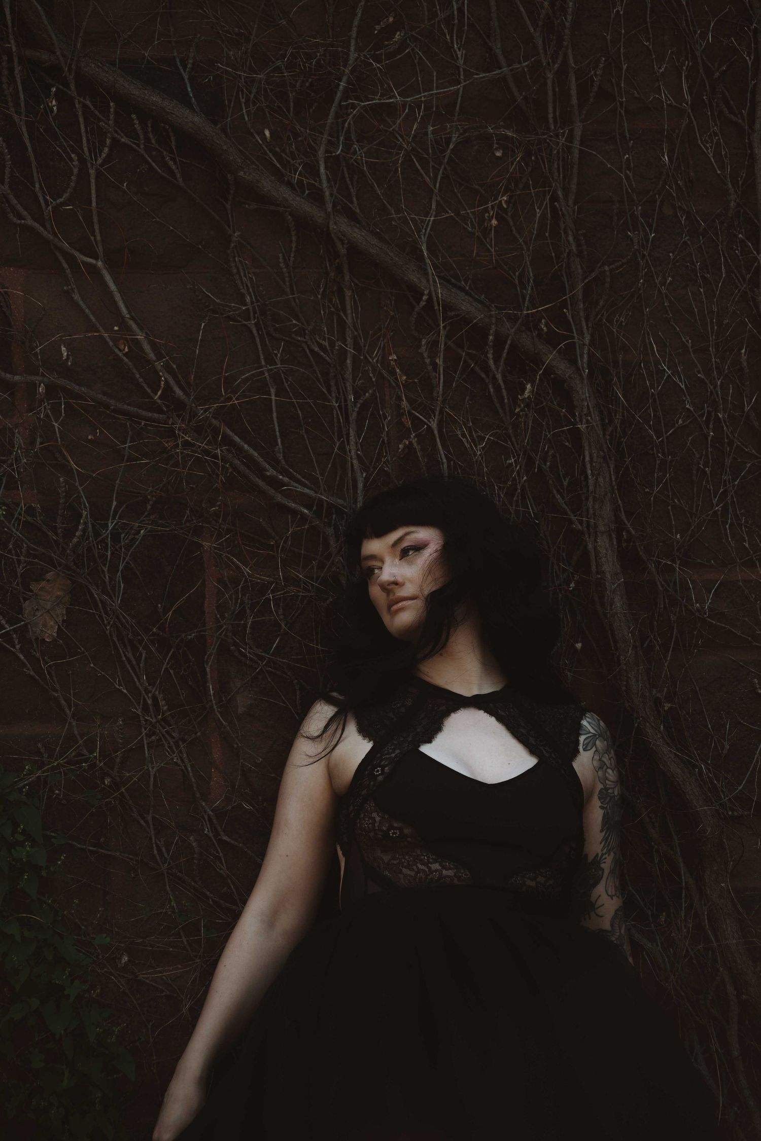 Woman in black lace dress with tattoo poses among bare dark branches in moody outdoor portrait photography.