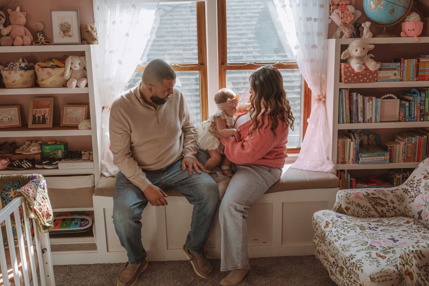 A family of three sits together in a cozy pink nursery, parents admiring their baby near a window.