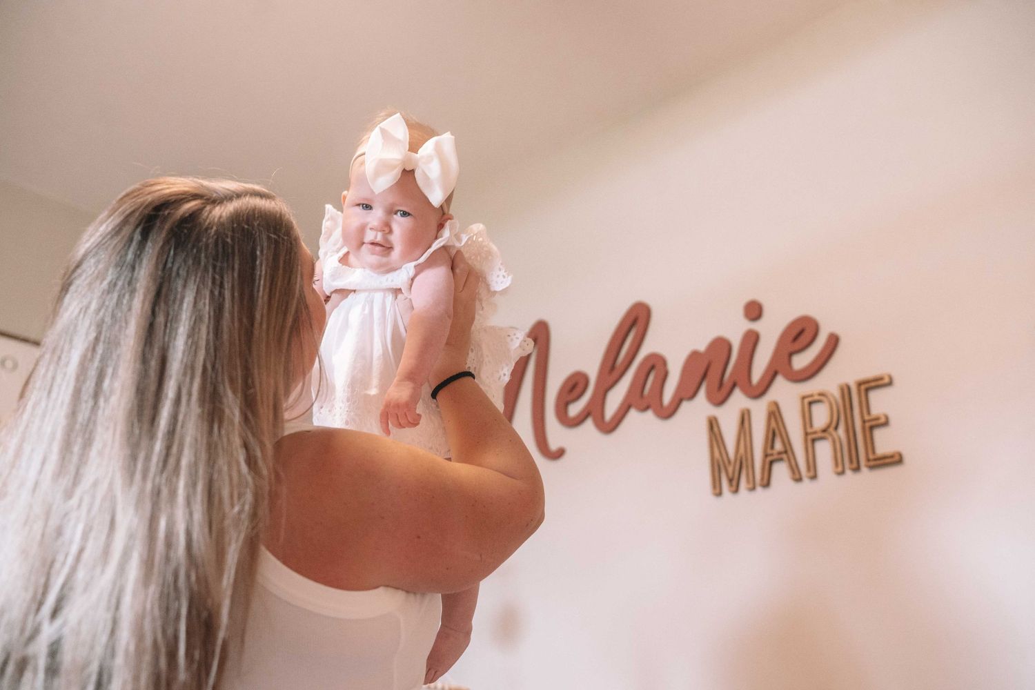 Mother lifting smiling baby girl named Melanie Marie, with wooden name sign on nursery wall.