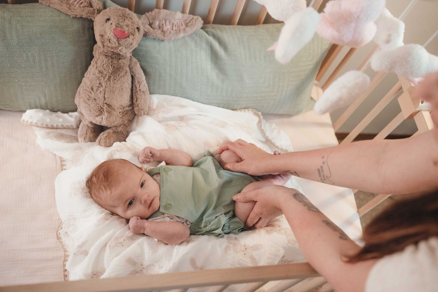 Baby in green onesie lying in white crib with stuffed bunny toy and pastel mobile above.