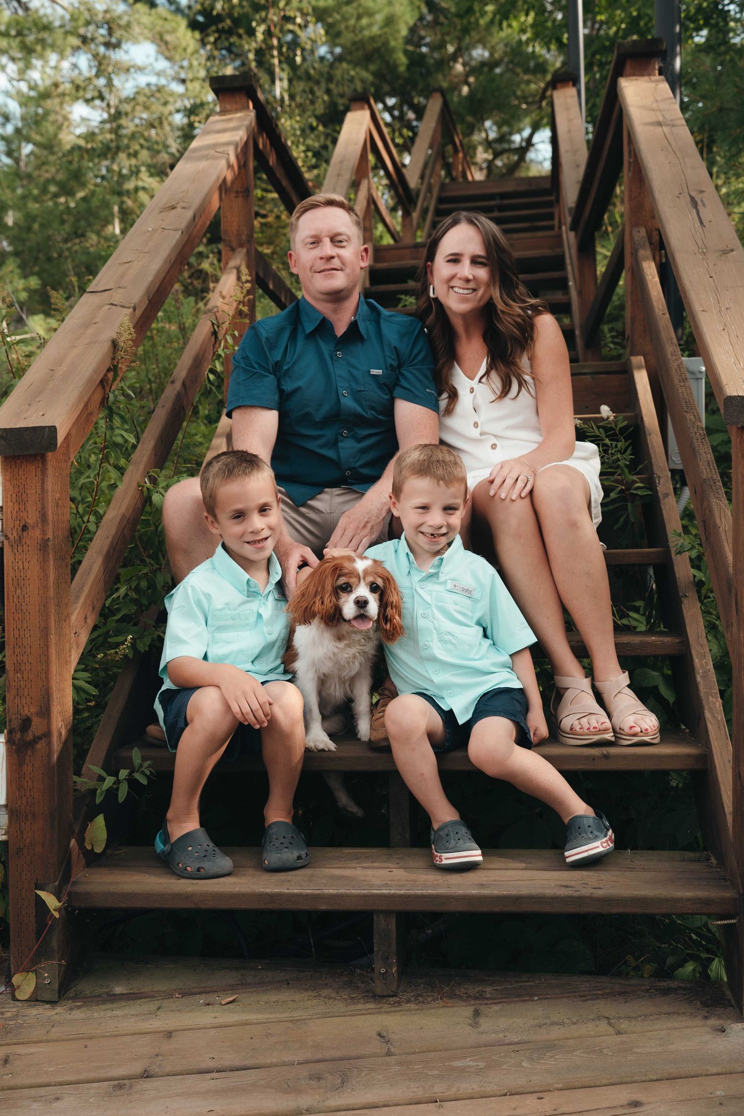 Family of four with a Cavalier King Charles Spaniel posing on wooden steps outdoors.