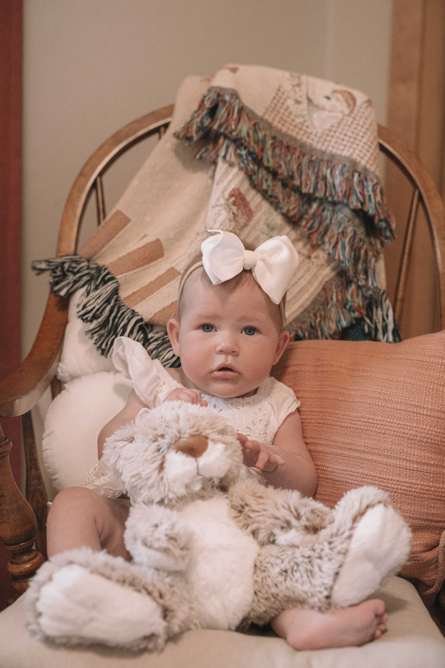Baby girl with white bow headband sits in rattan chair holding stuffed bunny toy, surrounded by cozy pillows and blankets.