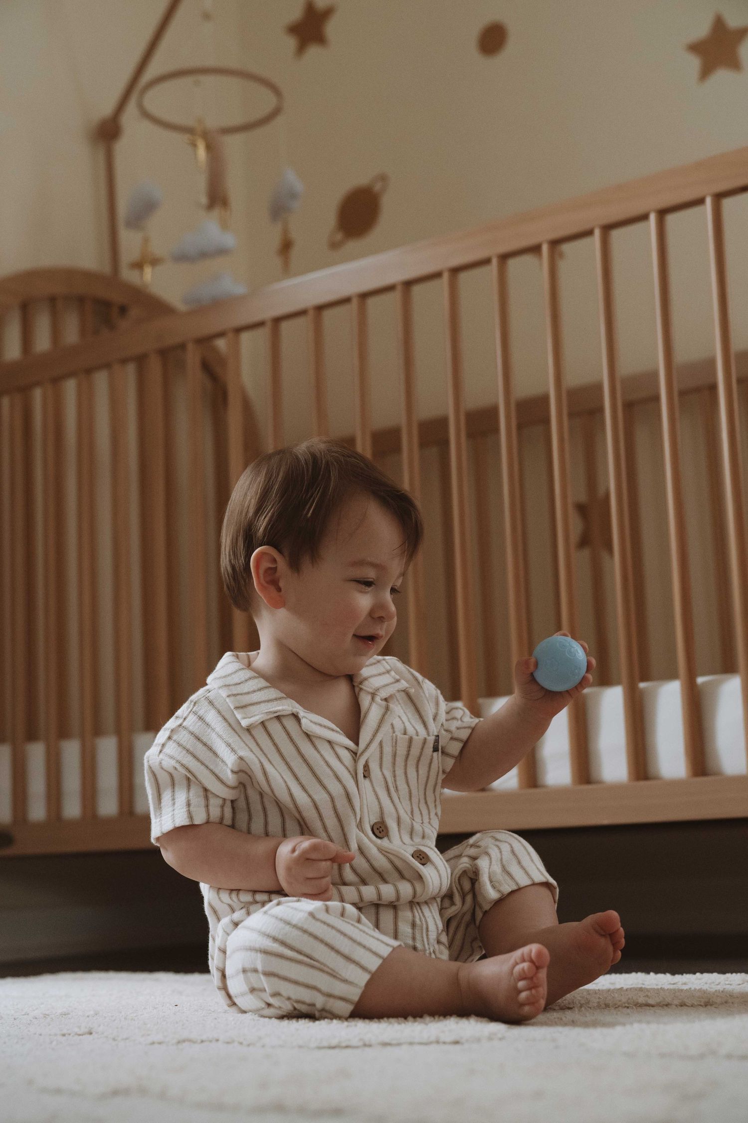 Smiling baby in striped pajamas sitting on carpet holding blue ball in front of wooden crib in nursery.