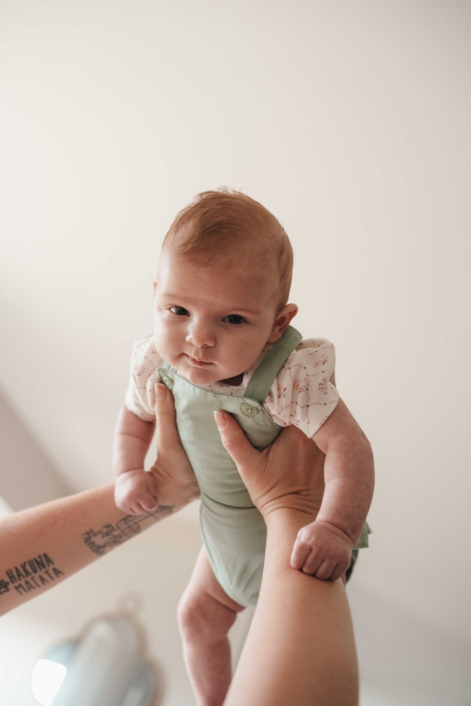 Adult hands with tattoo lifting a cute baby wearing a green romper and floral onesie against a white background.