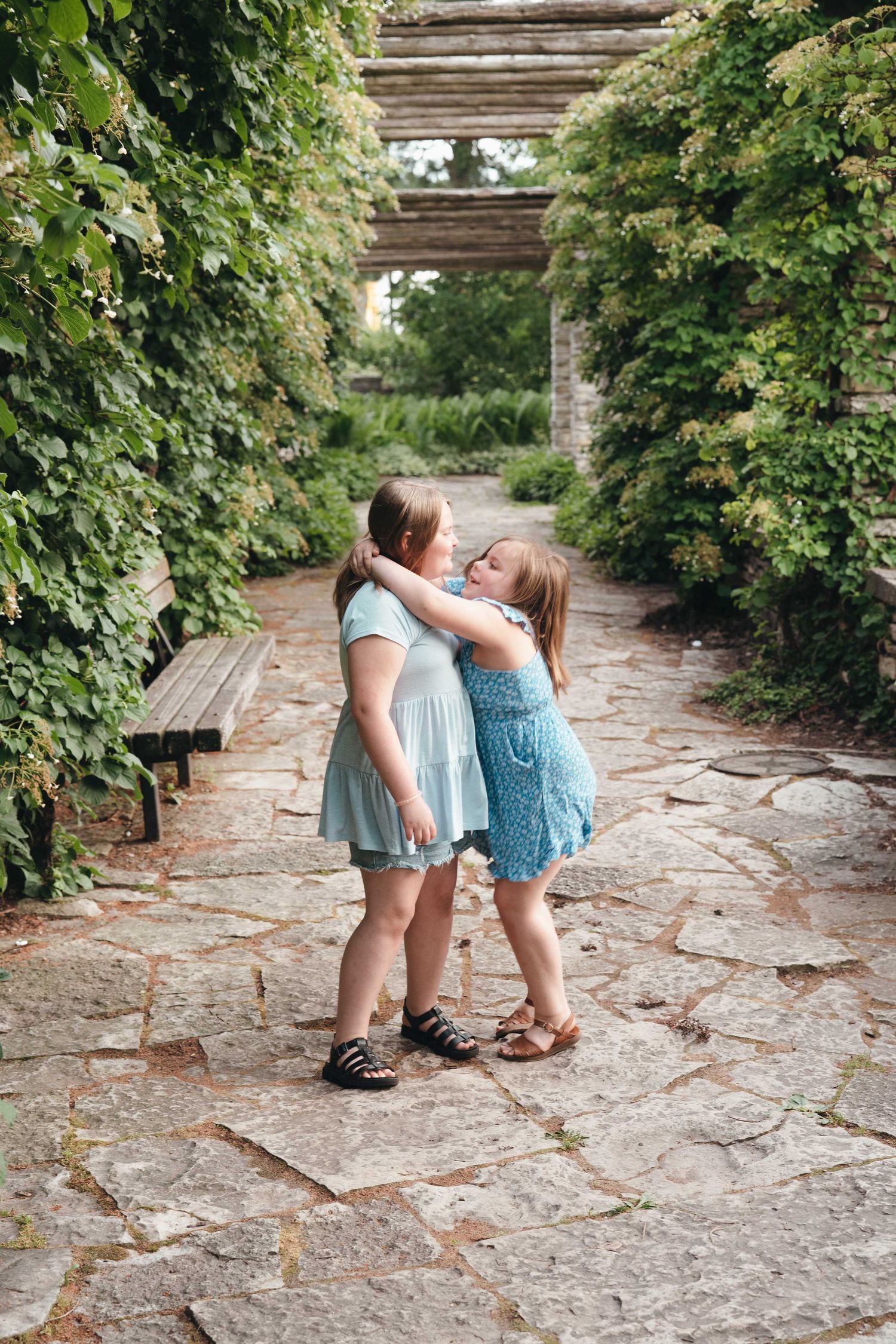 Two young girls in blue dresses hugging on a stone path surrounded by lush green garden pergola.