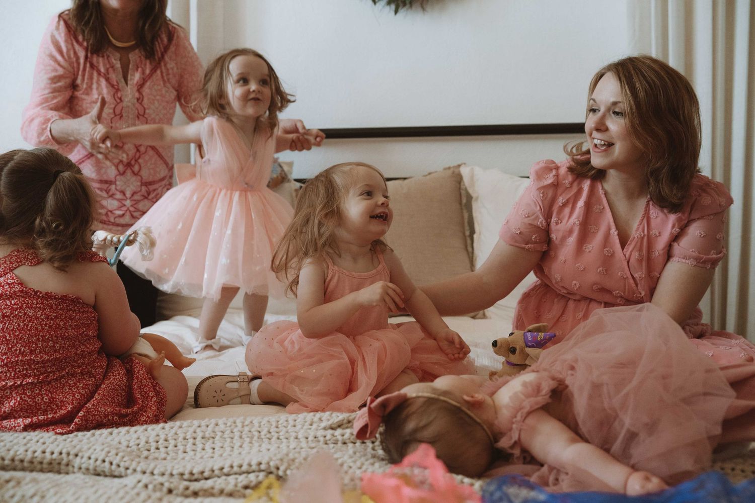 A woman in a pink dress sits on the floor laughing with three young girls in pink dresses in a bright living room.