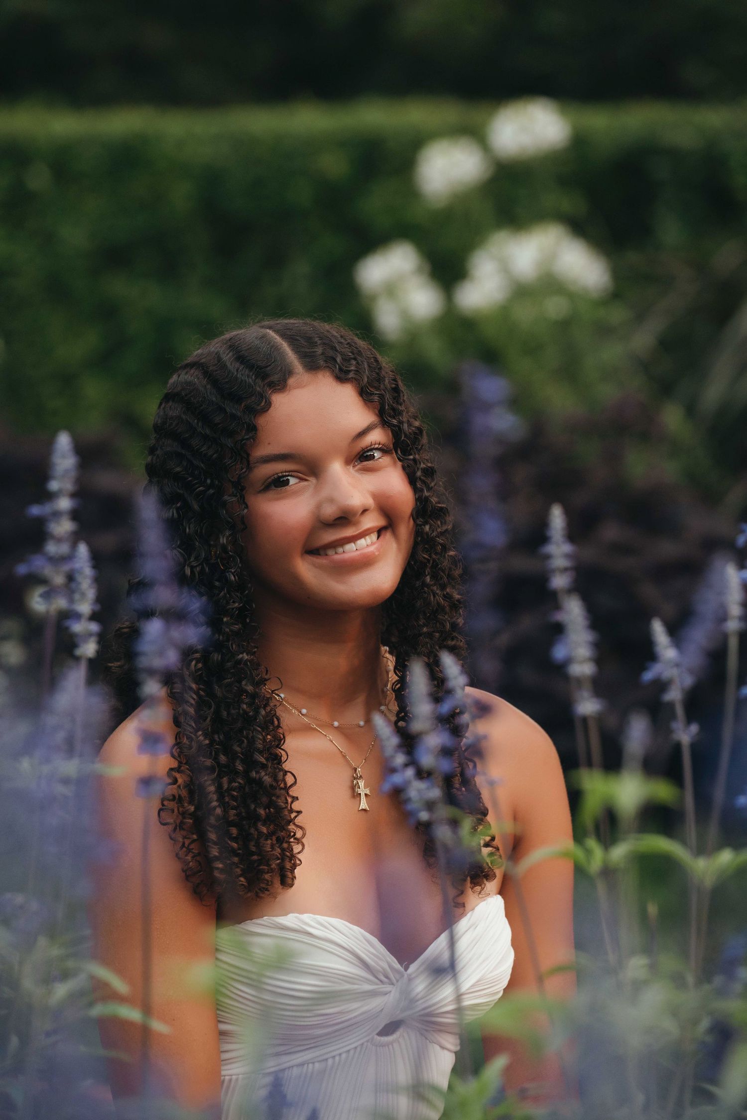 Young woman with curly hair smiling in a garden surrounded by purple lavender flowers, wearing a white dress and gold necklace.