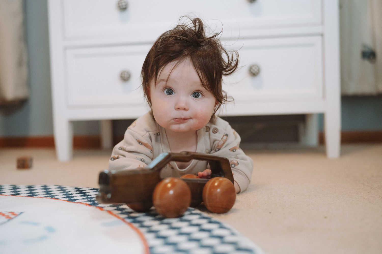 Cute baby with messy dark hair crawling on carpet playing with a wooden toy car in a bright nursery room.
