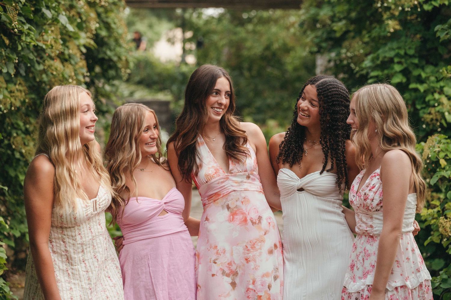Five women in floral and pastel summer dresses laughing together in a lush green garden setting.