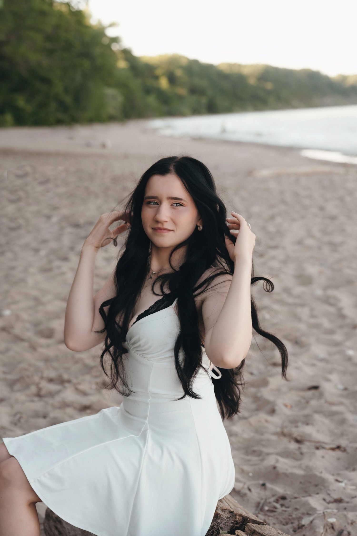 Young woman with long dark hair in white dress sitting on beach log, posing at sunset with trees in background.