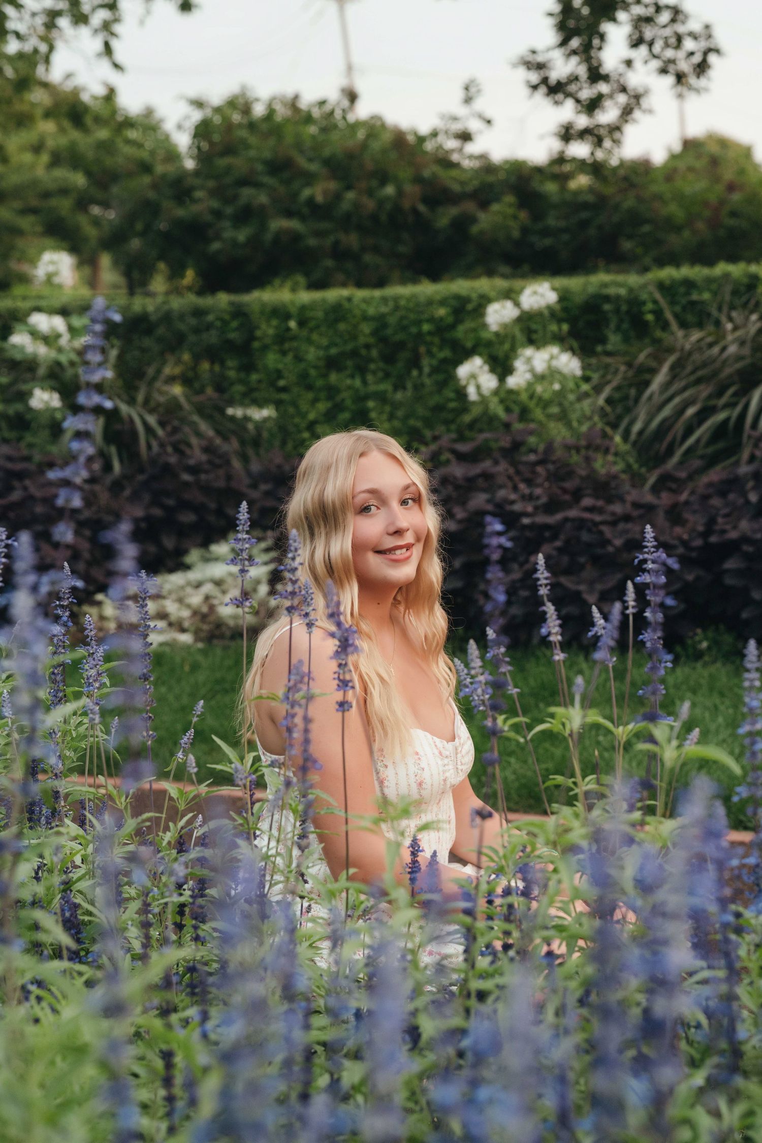 Young blonde woman sitting among purple lavender flowers in a lush green garden, smiling softly.