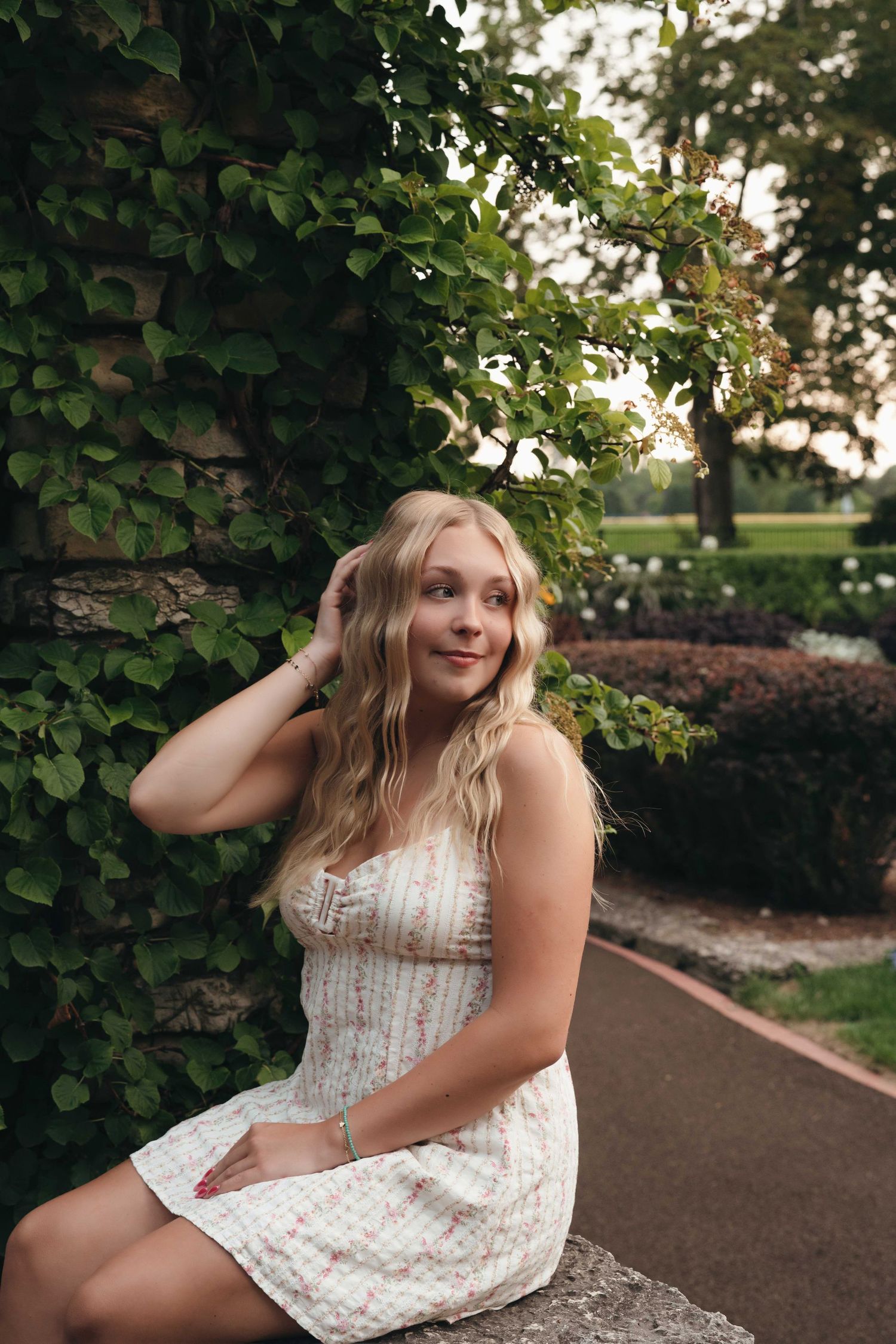 Young blonde woman in floral sundress sitting by ivy-covered stone wall in a lush garden park setting.