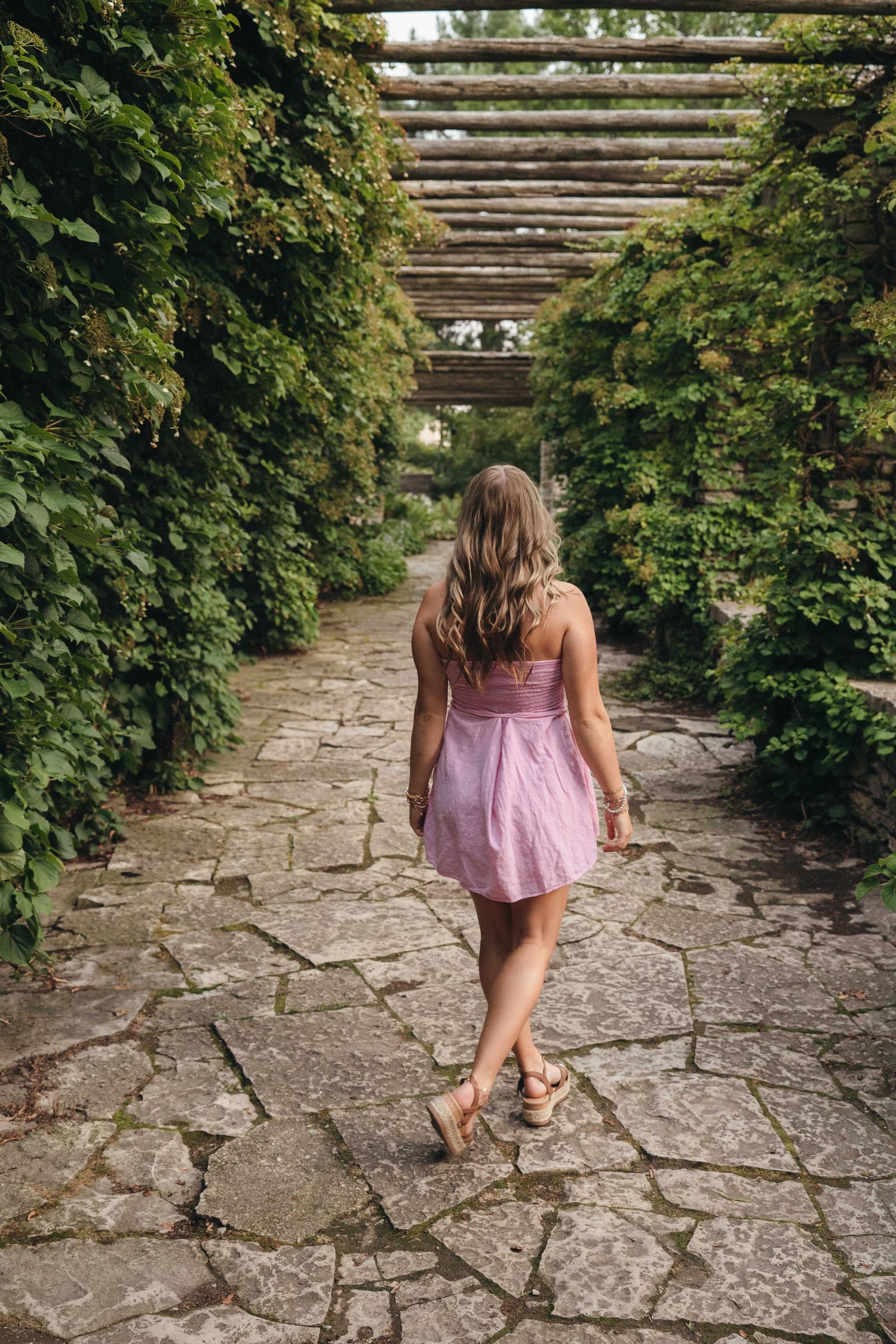Woman in pink dress walking along a stone path through a lush green pergola garden tunnel.