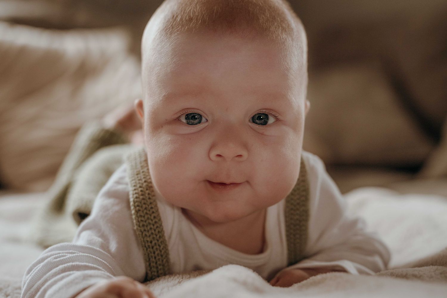 Adorable baby with blue eyes doing tummy time on a soft bed, wearing a white onesie and green suspenders.