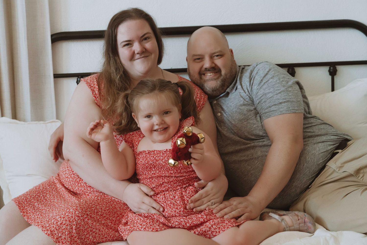 Happy family of three smiling together on a bed, parents with young daughter in red dress.