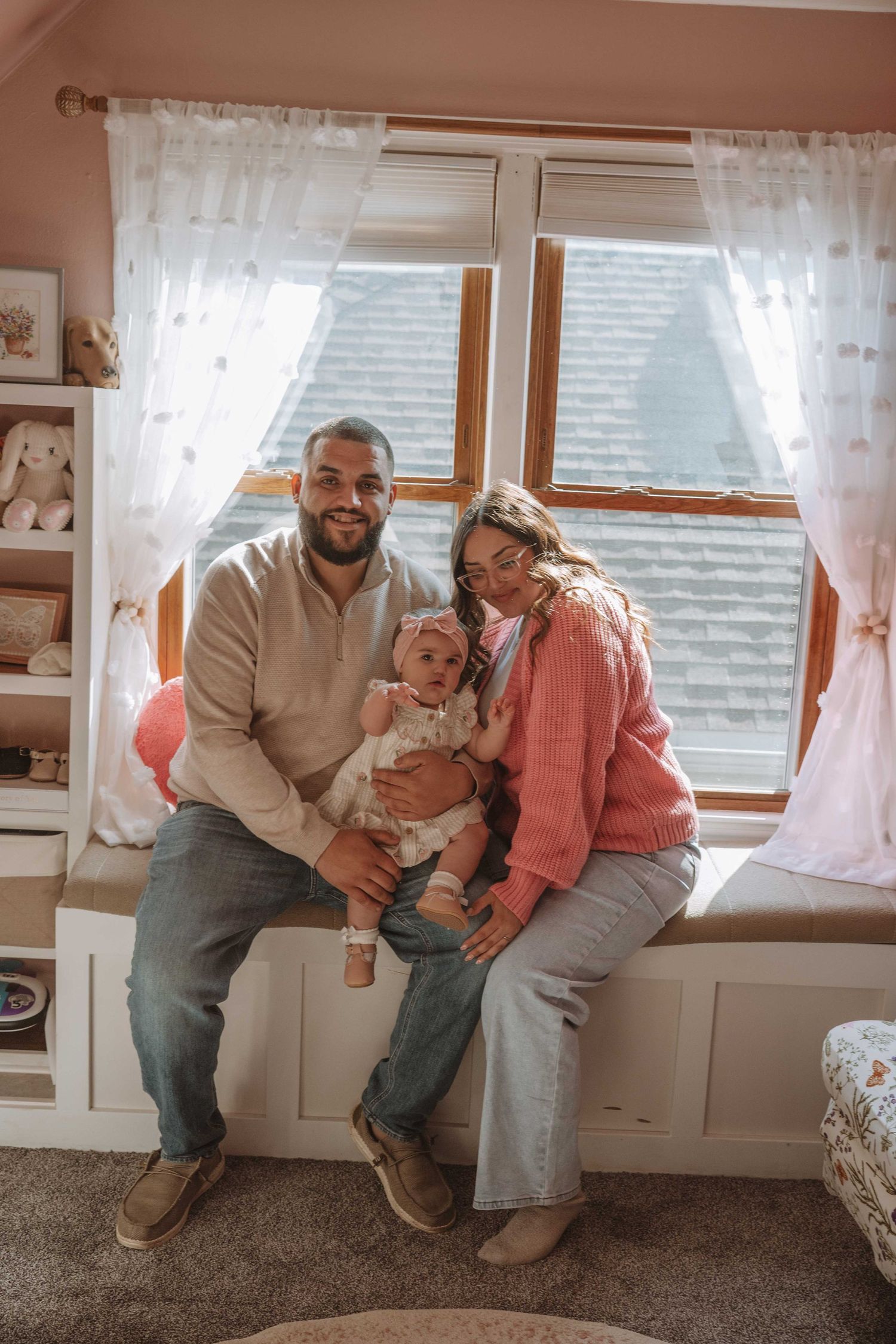 A happy young family with a baby sits on a window bench in a cozy pink nursery room.