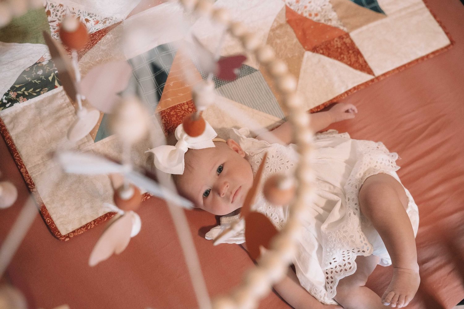 Baby girl in white lace dress lying on terracotta surface with colorful quilt and decorative branches in foreground.
