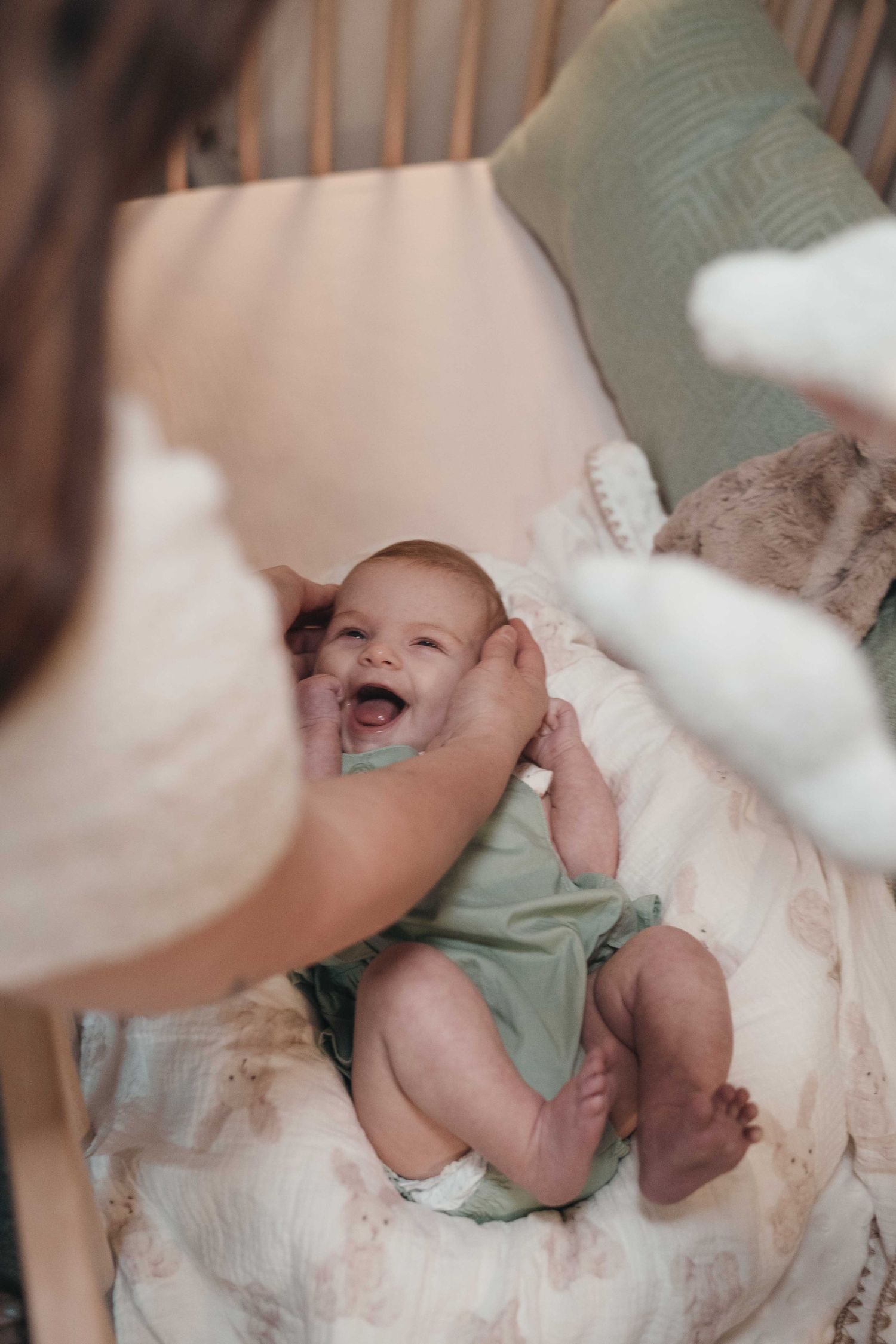 A laughing baby in a green onesie lies on soft cushions while a caregiver playfully interacts with them.