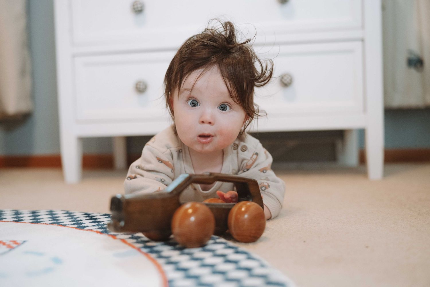 Cute baby with dark hair playing with a wooden toy car on the floor near a white dresser.