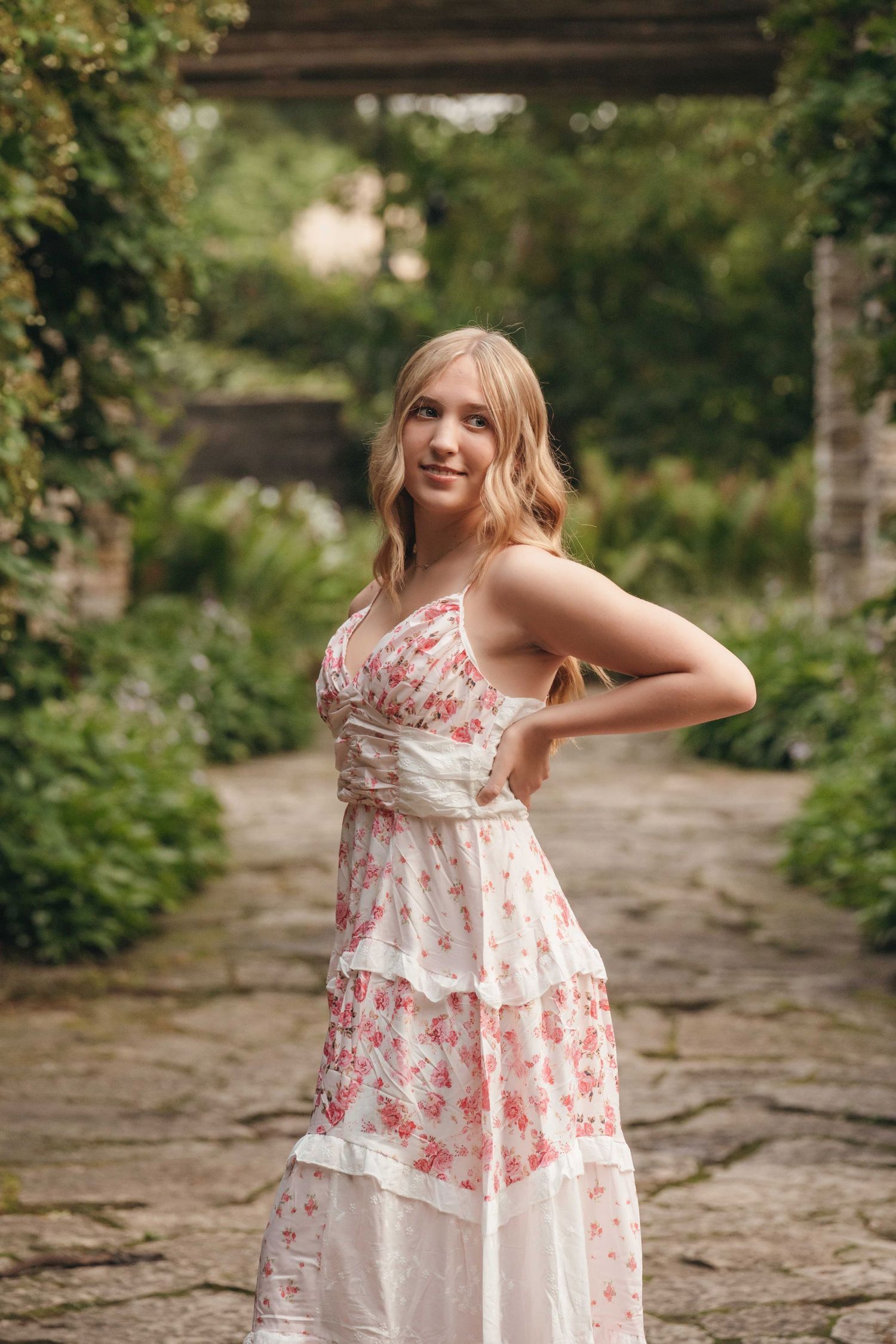 Young blonde woman in floral sundress posing in a lush garden pathway with stone walkway.