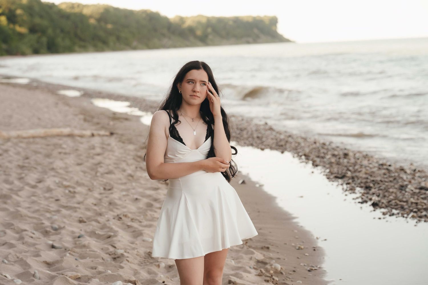 Young woman in white dress walking on sandy beach near calm water with green trees in background.