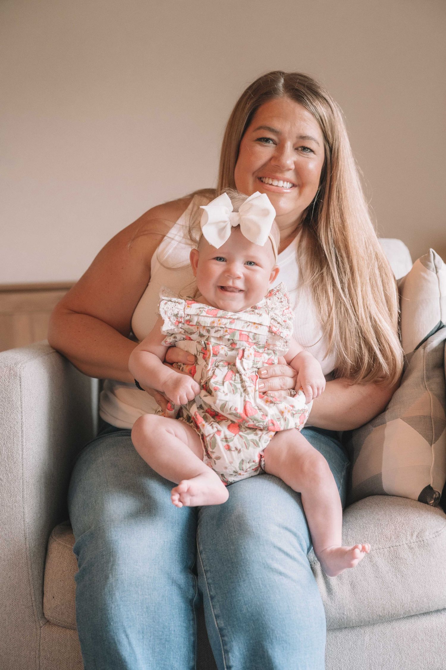 Smiling woman sitting in gray chair holding happy baby girl in floral romper with white bow headband.