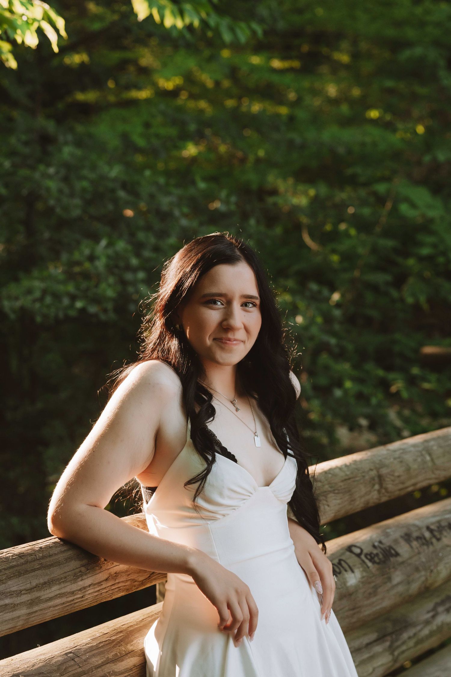 Young woman in white dress leaning on wooden fence, smiling softly with lush green trees in background.