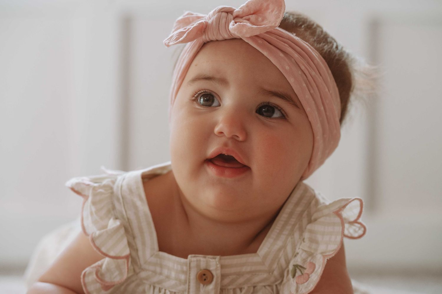 Adorable baby girl wearing pink bow headband and white ruffled outfit looking up curiously.