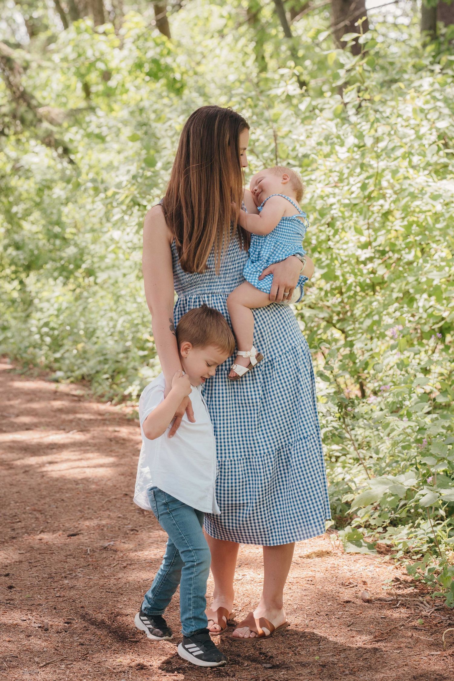 A mother in a blue dress holds her baby while her toddler hugs her on a sunny forest path.