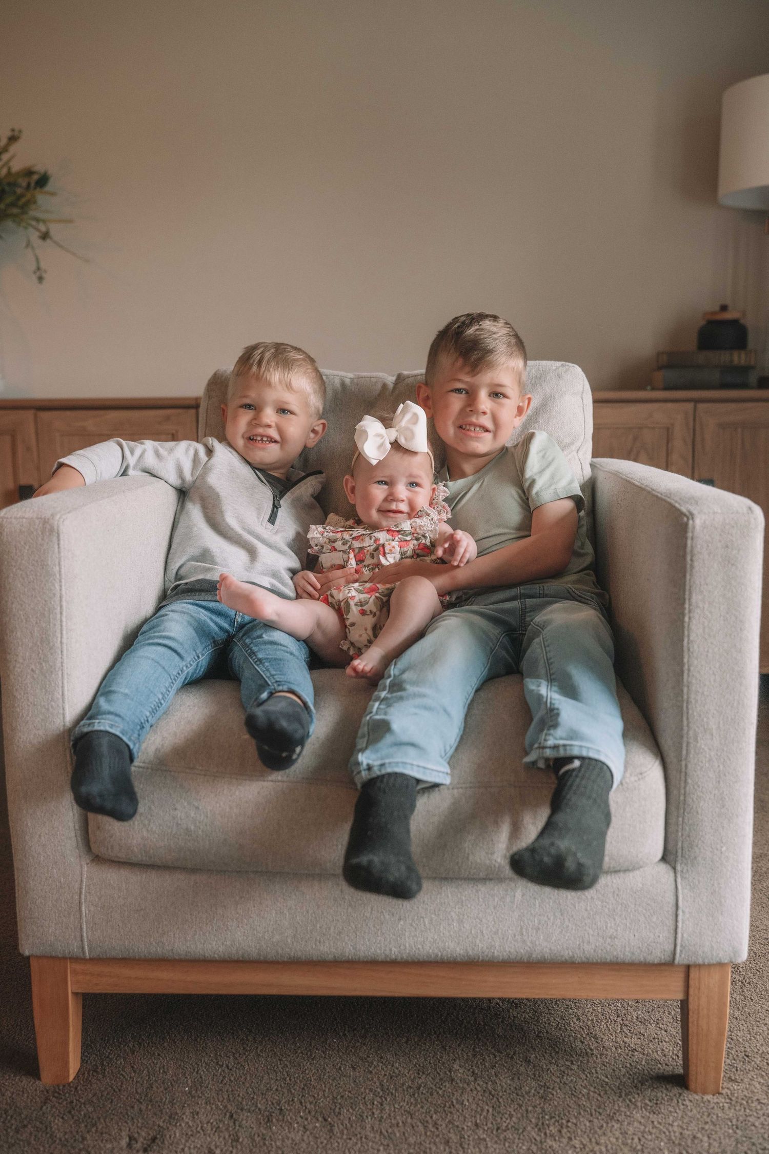 Two young boys and a baby girl in floral outfit sit together smiling on a beige armchair in a cozy living room.