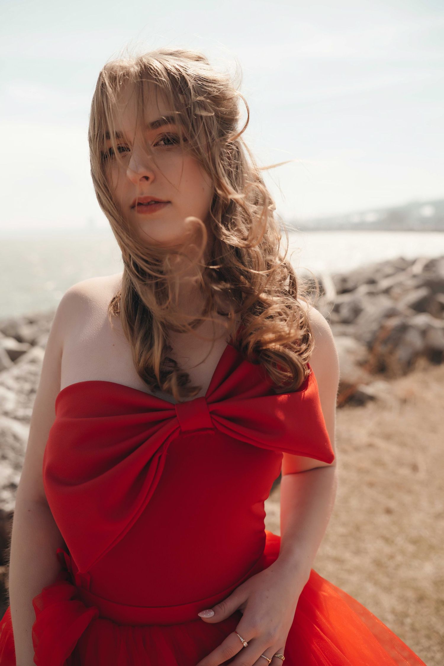 A person in a flowing red dress poses dramatically against a rocky beach backdrop on a sunny day.