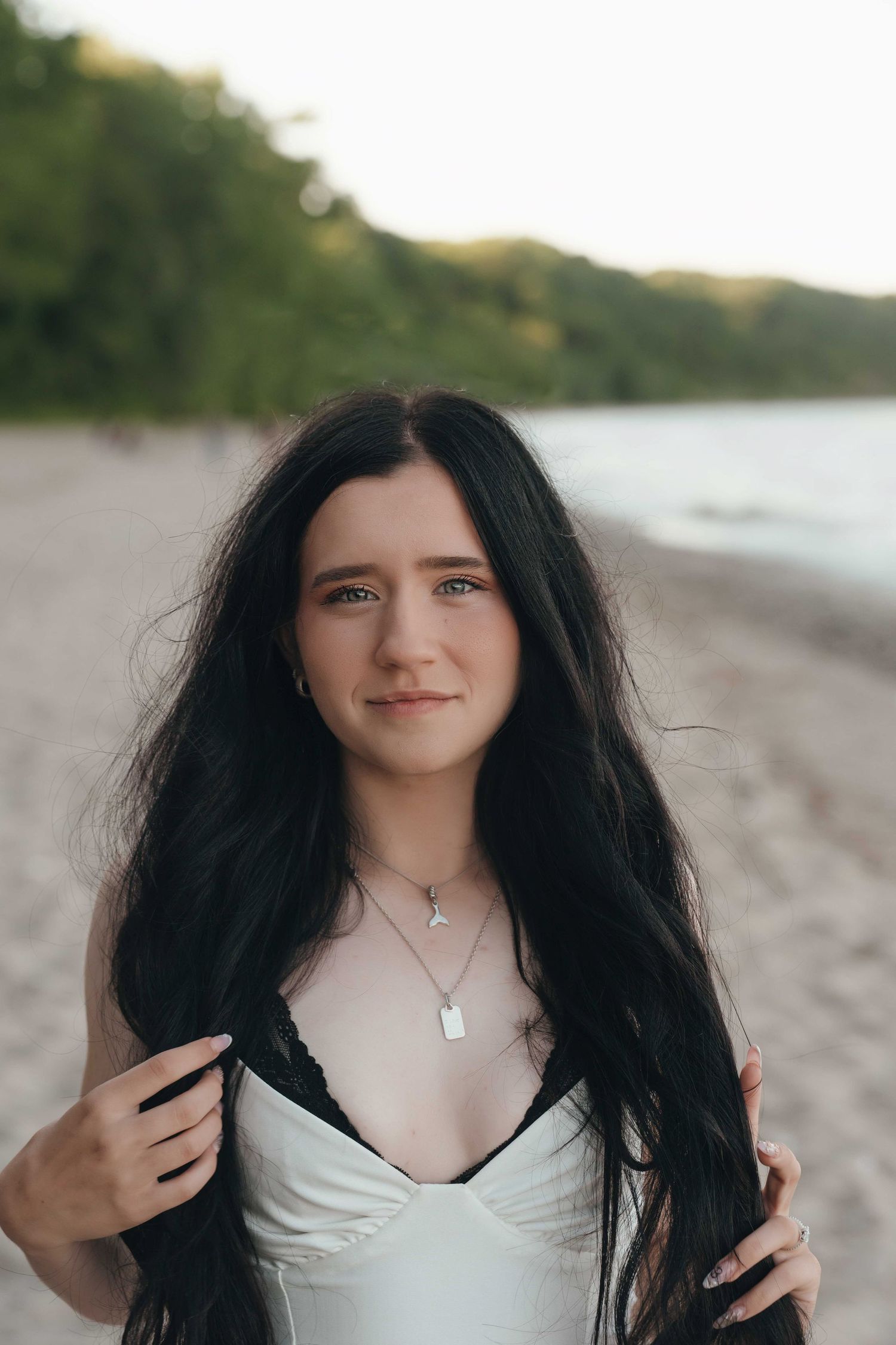 Young woman with long dark hair wearing a white top and necklace, standing on a sandy beach at sunset.