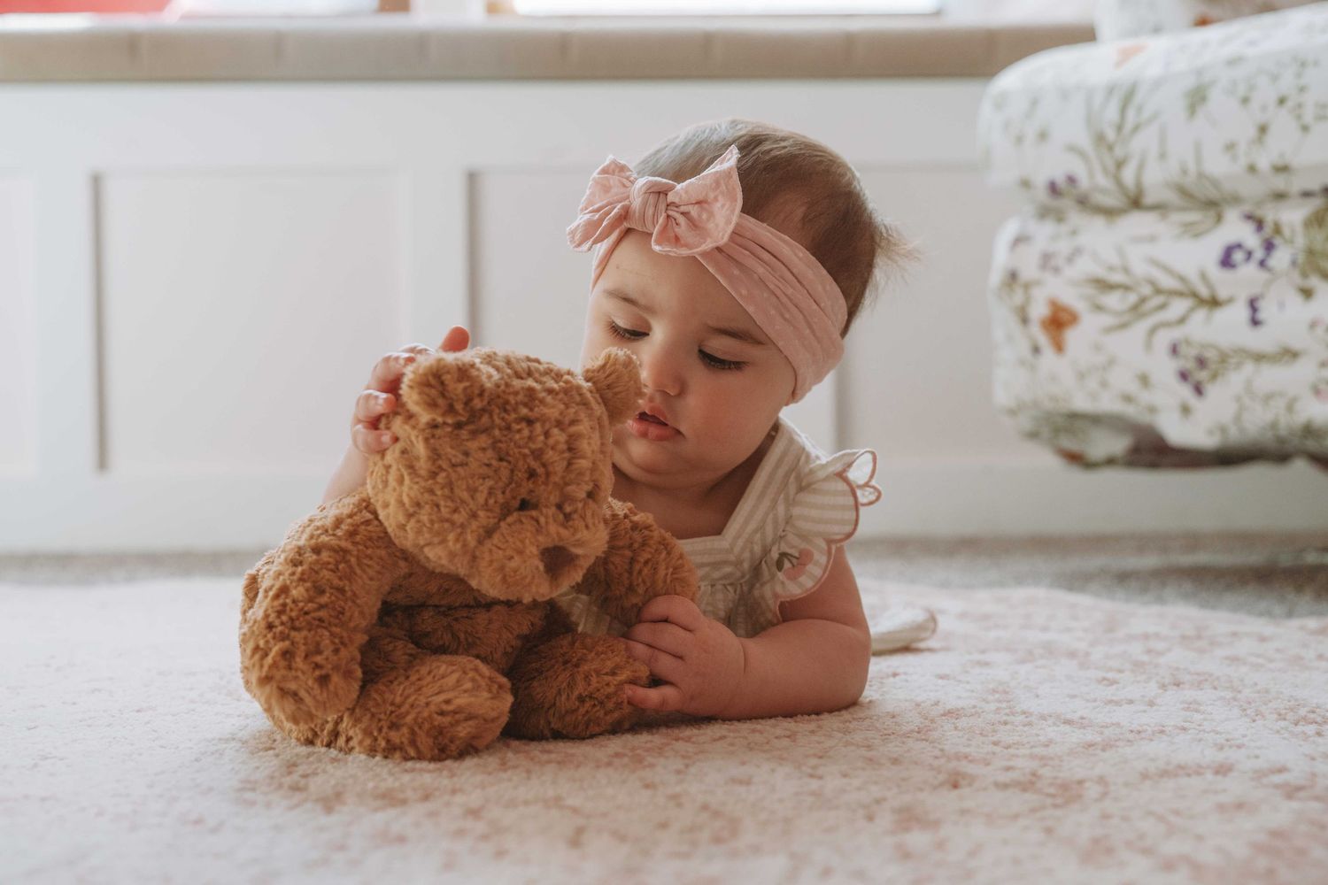 Adorable baby girl in a white dress with a pink bow headband cuddles a brown teddy bear on a soft carpet.