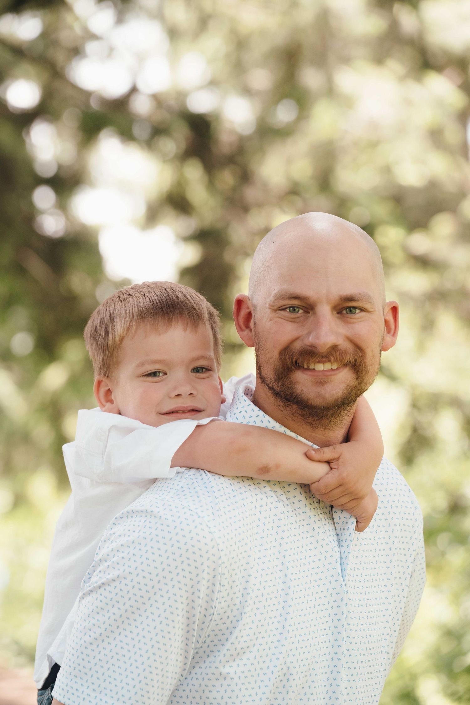 Smiling bald man with beard holding young blond boy outdoors, both wearing light blue shirts in a sunny park setting.