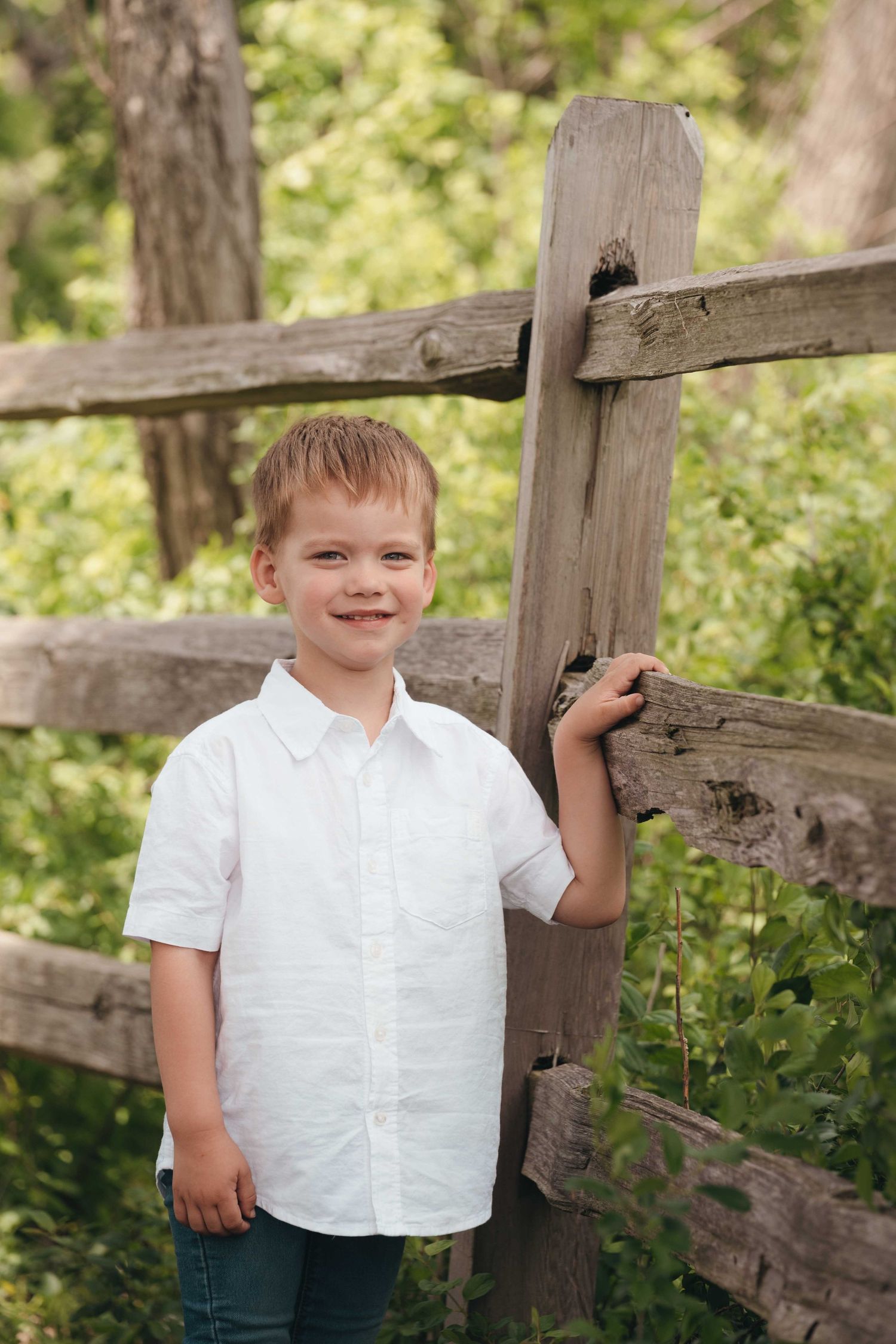 A smiling young boy in a white button-up shirt leans against a rustic wooden fence surrounded by lush green foliage.